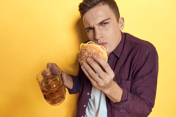 Homem alegre com uma caneca de cerveja e um hambúrguer em suas mãos dieta comida estilo de vida fundo amarelo — Fotografia de Stock