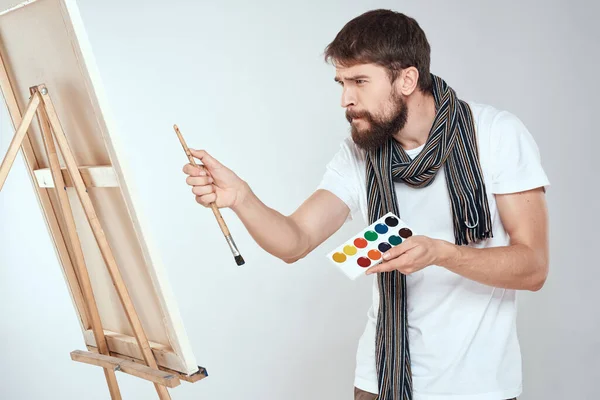 A man artist paints on an easel a brush and paint in the hands of a hobby creativity light background — Stock Photo, Image
