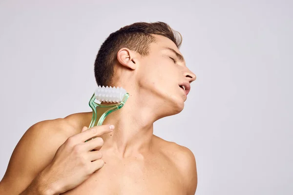 Un uomo nudo con un massaggiatore corpo su uno sfondo chiaro inclinato la testa al lato della pelle pulita — Foto Stock