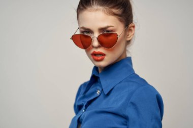 Güneş gözlüklü modaya uygun bir kadın ve elleri açık arka planda beliren mavi bir gömlek.