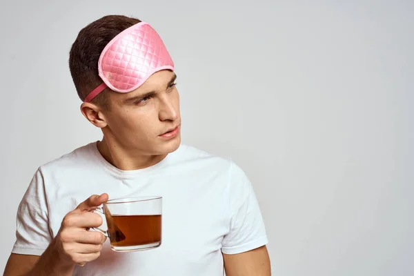 Bell'uomo con maschera rosa del sonno e con una tazza di tè in mano si tira su su su su su uno sfondo leggero — Foto Stock