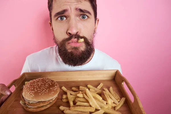 Muž s tácem s jídlem v ruce hamburger hranolky a rychlé občerstvení kalorie růžové pozadí portrét close-up — Stock fotografie