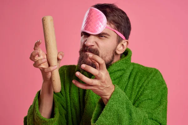 Retrato de um homem em uma máscara de sono rosa e um rolo de madeira emoções modelo de irritabilidade roupão verde — Fotografia de Stock