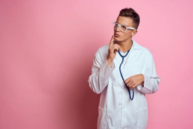 Pembe bir arka planda steteskop ve yüzünde gözlük olan tıbbi önlüklü bir erkek doktor.