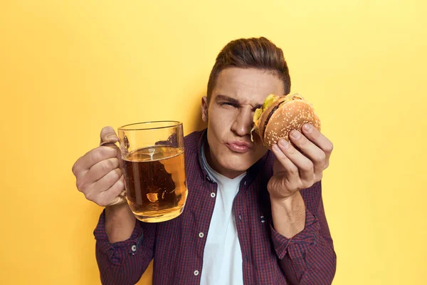 맥주 한 모금을 들고 손에 햄버거를 든 쾌활 한 남자가 노란 배경의 음식을 먹고 있다 — 스톡 사진