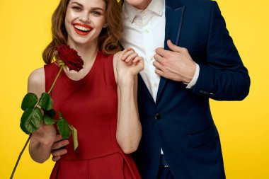 Romantik bir adam sarı arka planda elinde gülle kırmızı elbiseli bir kadına sarılır.