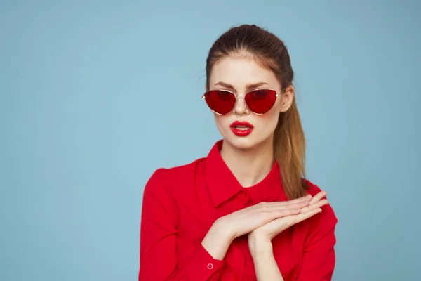 Mujer con camisa roja sobre fondo azul sostiene las manos cerca de la cara y gafas de sol labios rojos modelo de maquillaje — Foto de Stock