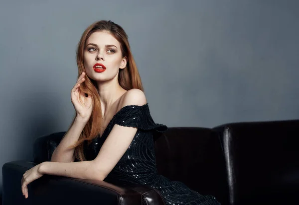 Mujer con encanto en vestido negro en un sofá de cuero modelo de maquillaje brillante — Foto de Stock
