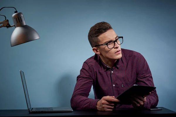 Um homem de óculos senta-se a uma mesa em um escritório com um laptop e uma lâmpada de mesa — Fotografia de Stock