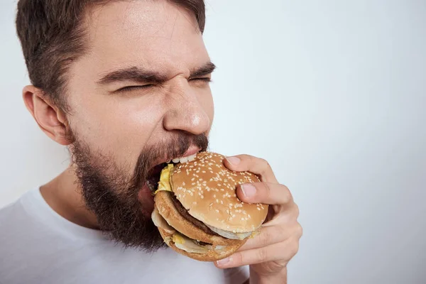 Um homem comendo um hambúrguer em um fundo leve em uma camiseta branca vista recortada close-up fome fast food — Fotografia de Stock