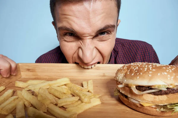 Hombre con paleta de madera comida rápida papas fritas hamburguesa hambre dieta alimentos fondo azul — Foto de Stock