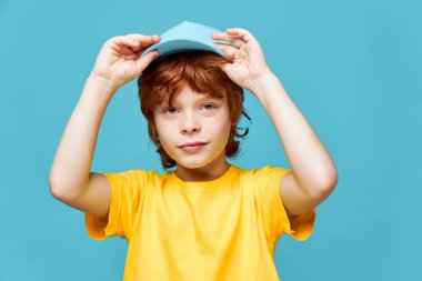 Kızıl saçlı çocuk kafasına şapka takıyor. Sarı tişörtü yakın plan çekilmiş. 