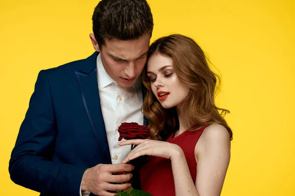 Romantický muž objímá ženu v červených šatech s růží v ruce na žlutém pozadí — Stock fotografie