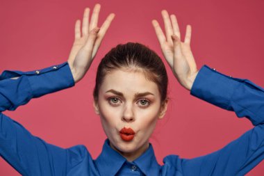 Yüzüne yakın elleri ve mavi gömleği olan, pembe arka planı olan enerjik bir kadın.