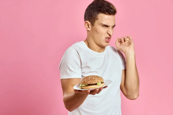 Emocional chico con hamburguesa en un plato y blanco camiseta rosa fondo recortado vista de las calorías de comida rápida — Foto de Stock