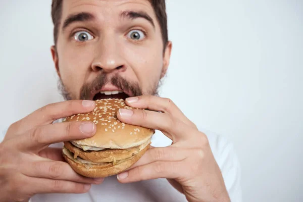 白いTシャツに身を包んだ男がハンバーガーを食べると、空腹のファーストフードが近くに見える — ストック写真