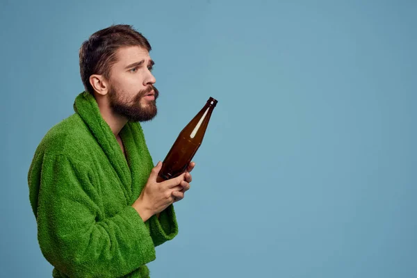 Пьяный мужчина в зеленом халате с бутылкой пива в руке на синем фоне алкоголя — стоковое фото
