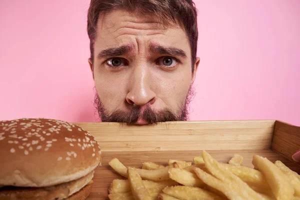 Muž s tácem s jídlem v ruce hamburger hranolky a rychlé občerstvení kalorie růžové pozadí portrét close-up — Stock fotografie