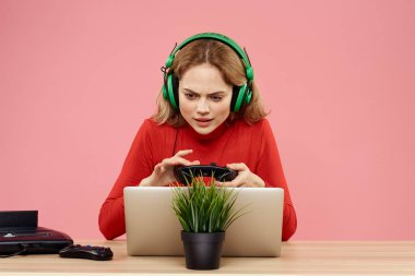 Kulaklık takan, dizüstü bilgisayarlı bir kadın masa başı eğlence sektöründe pembe arka planda oturuyor.