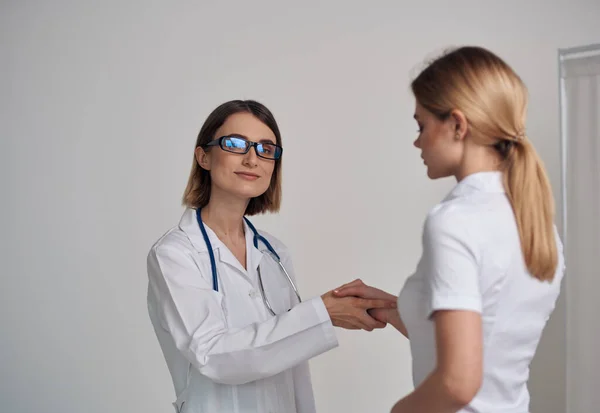 Kvinna läkare i en medicinsk klänning skakar hand med en patient i en vit t-shirt på en lätt bakgrund — Stockfoto