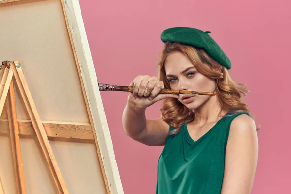 Kvinna konstnär grön basker staffli borste rita lektioner konst rosa bakgrund — Stockfoto