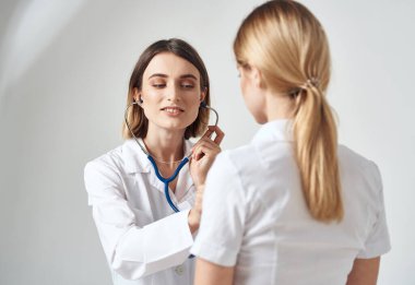 Steteskoplu bir doktor beyaz tişörtlü bir kadını muayene ediyor.