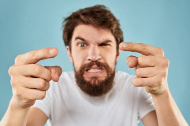 Beyaz tişörtlü sakallı adam. Duyguları, elleri ve yüz ifadesinden memnun olmayan mavi arka plan.