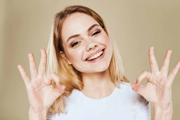 Mulher loira alegre em um branco gestos camiseta com suas emoções da mão fundo bege — Fotografia de Stock