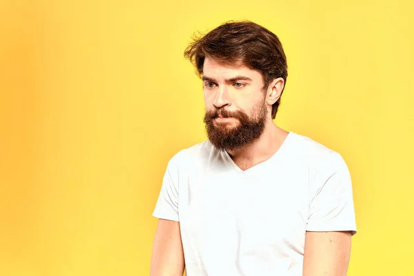 Barbudo hombre en una camiseta blanca gestos con las manos emociones estudio fondo amarillo —  Fotos de Stock