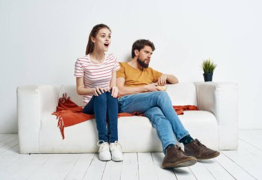 Beyaz bir kanepede bir adam ve bir kadın parlak bir odada bir tabak patlamış mısır ve bir saksıda bir çiçek.