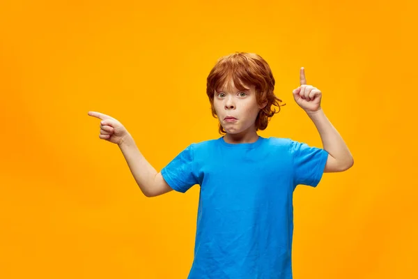 Roztomilý zrzek chlapec ukazuje prsty v různých směrech modré tričko kopírovat prostor. — Stock fotografie