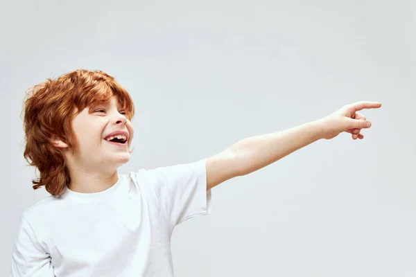 Wesoły rudowłosy dziecięcy gest z jego ręka wskazujący na bok biały T-shirt Copy Space — Zdjęcie stockowe