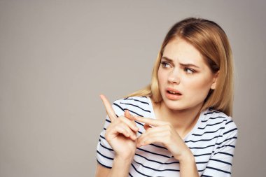 Yüzünde hoşnutsuzluk ifadesi olan bir kadın, elleri ve tişörtü gri arka planıyla el kol hareketi yapıyor.