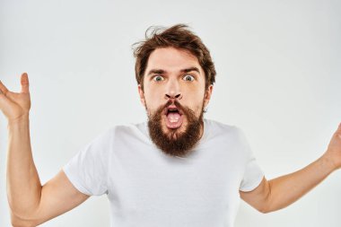Beyaz tişörtlü duygusal sakallı adam elleri açık arka planda jestler yapıyor.
