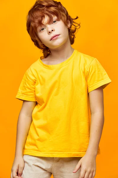 O menino ruivo em um fundo amarelo em uma camiseta e calças inclinou a cabeça — Fotografia de Stock