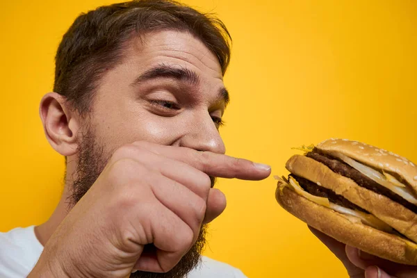 Hombre con hamburguesa en manos de comida rápida dieta blanca camiseta primer plano amarillo fondo — Foto de Stock