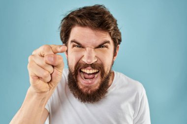 Beyaz tişörtlü sakallı adam. Duyguları, elleri ve yüz ifadesinden memnun olmayan mavi arka plan.