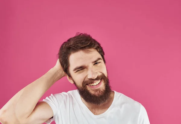 Felice ragazzo in una t-shirt bianca su uno sfondo rosa con una barba spessa sta gesticolando con le mani — Foto Stock