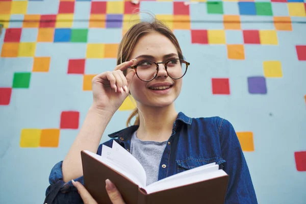 Студентка в рубашке и очках держит книгу в руках на улице возле граффити на стене — стоковое фото