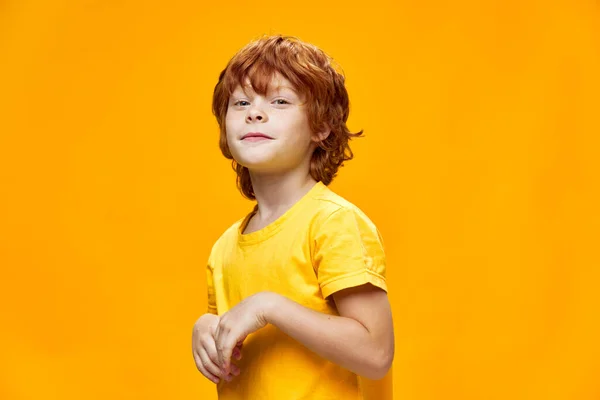 Um rapaz modesto com cabelo ruivo e uma t-shirt amarela segura as mãos — Fotografia de Stock