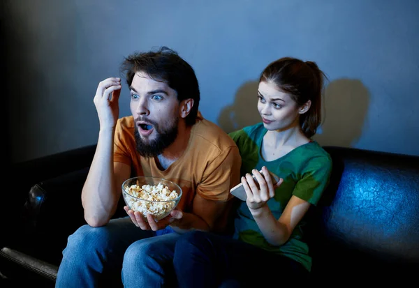 a man and a frightened woman watching a movie in the evening on the couch with a plate of popcor
