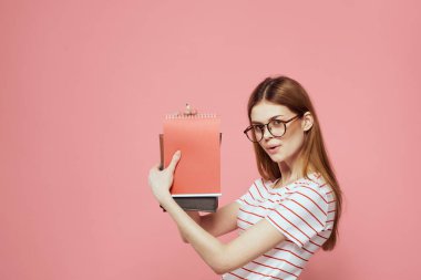 Pembe arka planda not defterleri olan, elleri gözlüklü, yüz çizgili tişört mankeniyle el kol hareketi yapan bir kız öğrenci.