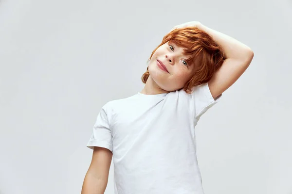 Glada rödhåriga pojke bugar huvudet åt ena sidan med handen på huvudet vit t-shirt — Stockfoto