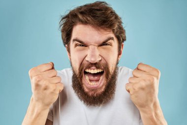 Beyaz tişörtlü sakallı adam. Duyguları, elleri ve yüz ifadesinden memnun olmayan mavi arka plan.