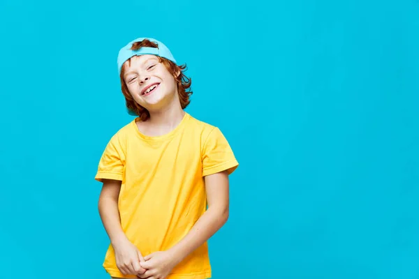 喜びの赤い髪の少年は彼の頭を傾けた片側ブルーキャップ黄色のTシャツ子供時代のスタジオ — ストック写真