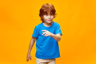 Neşeli kızıl saçlı çocuk elleriyle işaret ediyor. Mavi tişört, sarı izole edilmiş arka plan. 