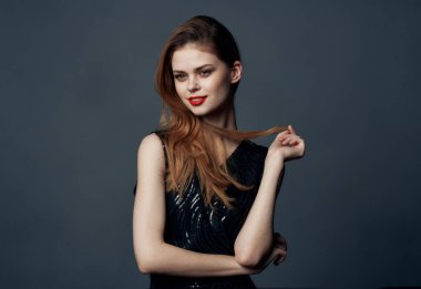 Siyah elbiseli, gri arka planda kızıl saçlı bir kadın elleriyle işaret ediyor.