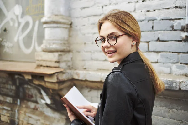 Gelukkige vrouw in een leren jas met een boek in de hand loopt door de straat in de stad — Stockfoto