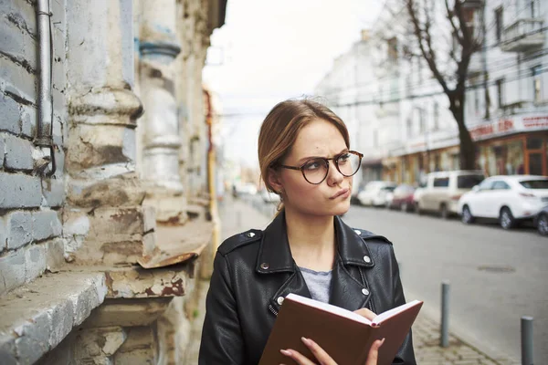 Femme heureuse dans une veste en cuir avec un livre à la main marche dans la rue dans la ville — Photo