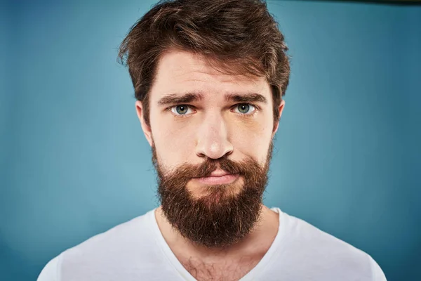 Bearded man displeased facial expression emotions close-up blue background white t-shirt